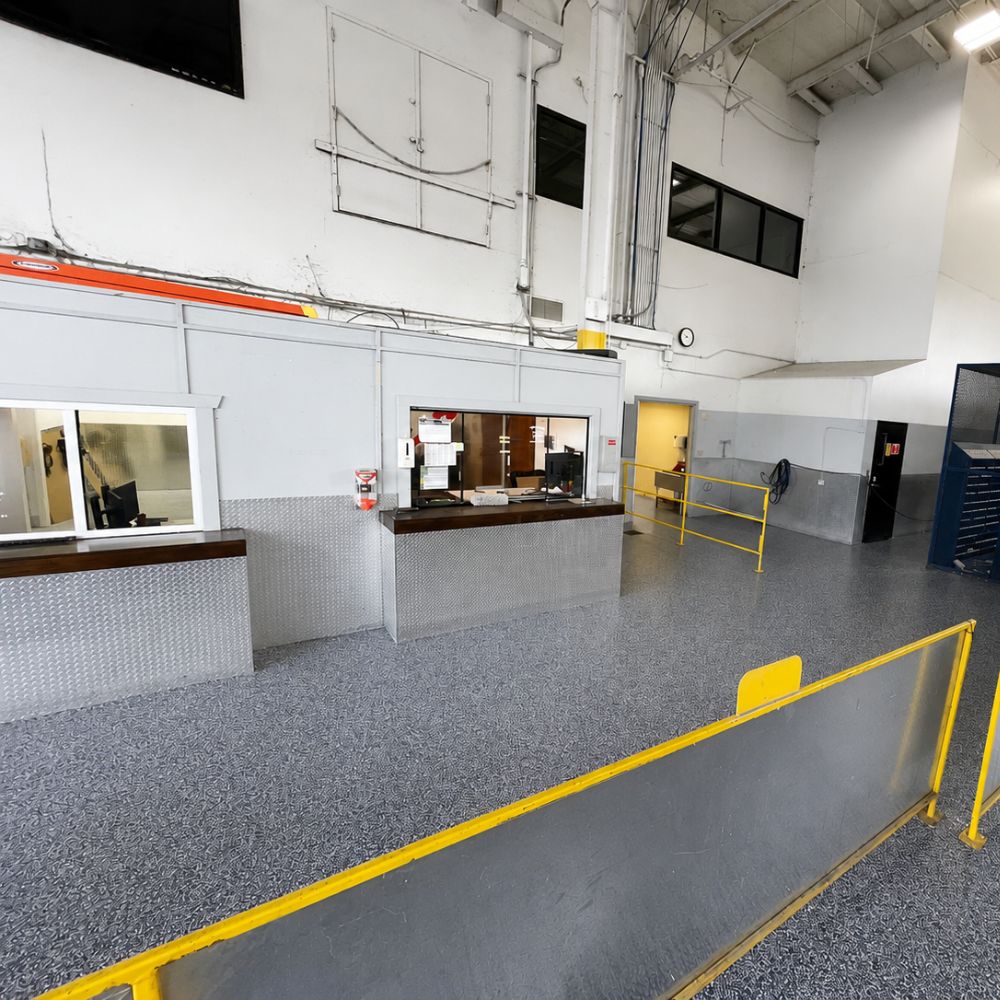 Industrial warehouse reception: a service counter with a window, diamond-plate walls, and yellow safety rails by a ramped walkway.