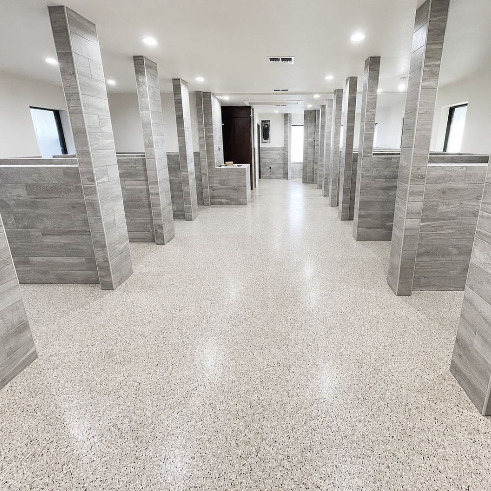 Long, well-lit public restroom or changing area with rows of grey tiled stalls on both sides and a speckled terrazzo floor.