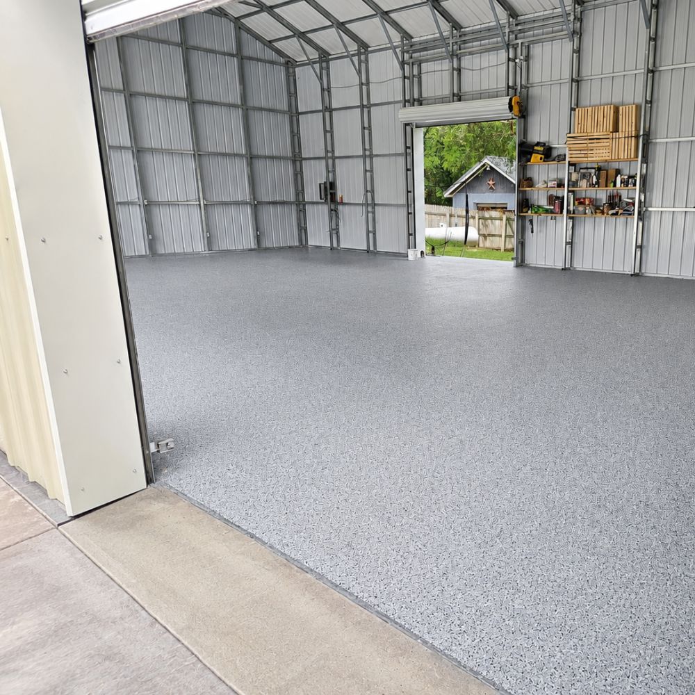 Empty metal workshop with speckled epoxy floor, shelves of tools on the right, and an open roll‑up door showing a yard outside.