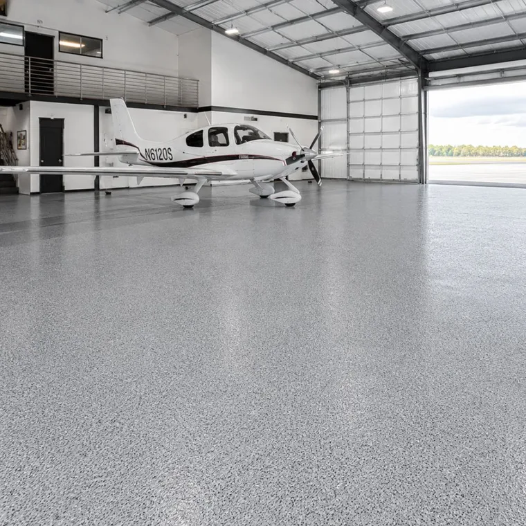 Single-engine airplane parked inside a hangar on a light gray flake polyaspartic floor, with the hangar door open to a runway