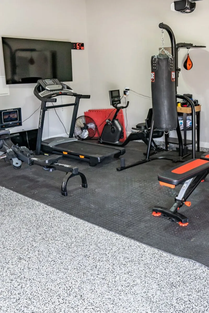 A home gym in a garage with a treadmill, a weight bench and a punching bag on a flake floor